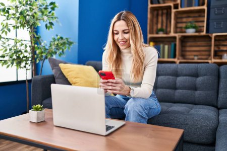 Téléchargez les photos : Jeune femme blonde utilisant un smartphone et un ordinateur portable à la maison - en image libre de droit