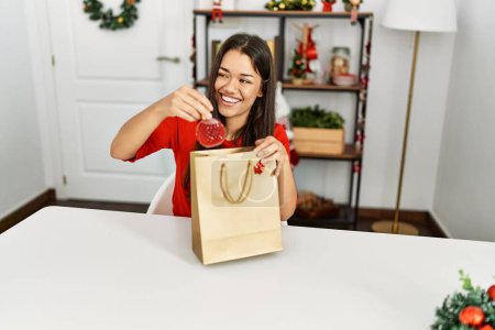 Téléchargez les photos : Jeune femme latine tenant parfum de sac cadeau assis par décor de Noël à la maison - en image libre de droit