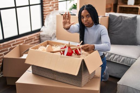 Téléchargez les photos : Femme afro-américaine déménageant dans une nouvelle maison boîtes d'ouverture avec la main ouverte faisant stop signe avec expression sérieuse et confiante, geste de défense - en image libre de droit