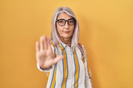 Photo for Middle age woman with grey hair standing over yellow background wearing glasses doing stop sing with palm of the hand. warning expression with negative and serious gesture on the face. - Royalty Free Image
