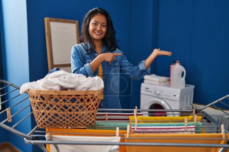 Foto de Joven mujer asiática colgando ropa en el tendedero sorprendido y sonriendo a la cámara mientras se presenta con la mano y señalando con el dedo. - Imagen libre de derechos