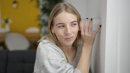 Téléchargez les photos : Young blonde woman listening through wall with glass at home - en image libre de droit