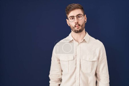 Photo for Hispanic man with beard standing over blue background making fish face with lips, crazy and comical gesture. funny expression. - Royalty Free Image