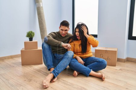 Téléchargez les photos : Jeune couple assis sur le sol à la nouvelle maison en regardant l'heure de la montre inquiet, peur de se faire tard - en image libre de droit