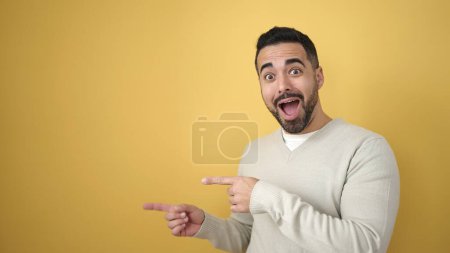 Téléchargez les photos : Jeune homme hispanique souriant confiant pointant avec les doigts sur le côté sur fond jaune isolé - en image libre de droit