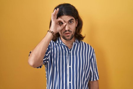 Photo for Hispanic man with long hair standing over yellow background doing ok gesture shocked with surprised face, eye looking through fingers. unbelieving expression. - Royalty Free Image