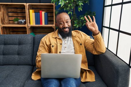 Foto de Hombre afroamericano usando el ordenador portátil en casa sentado en el sofá haciendo signo ok con los dedos, sonriendo gesto amistoso excelente símbolo - Imagen libre de derechos