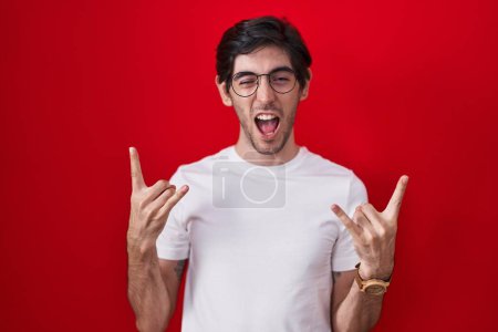 Téléchargez les photos : Jeune homme hispanique debout sur fond rouge criant avec une expression folle faire symbole rock avec les mains en l'air. star de la musique. concept de musique lourde. - en image libre de droit