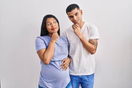 Photo for Young hispanic couple expecting a baby standing over background with hand on chin thinking about question, pensive expression. smiling with thoughtful face. doubt concept. - Royalty Free Image