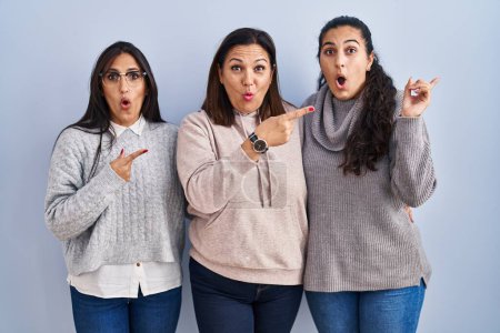 Photo for Mother and two daughters standing over blue background surprised pointing with finger to the side, open mouth amazed expression. - Royalty Free Image