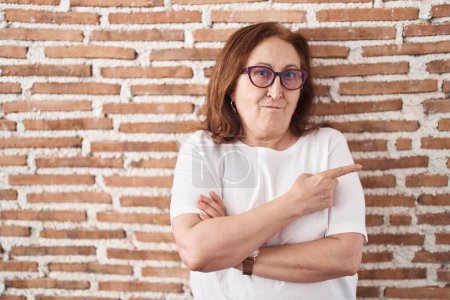 Téléchargez les photos : Senior woman with glasses standing over bricks wall pointing with hand finger to the side showing advertisement, serious and calm face - en image libre de droit