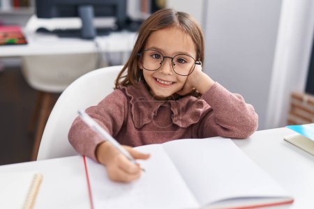 Téléchargez les photos : Adorable hispanic girl student sitting on table doing homework at classroom - en image libre de droit