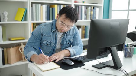 Foto de Young chinese man student using computer writing on notebook at library university - Imagen libre de derechos