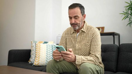 Téléchargez les photos : Homme d'âge moyen utilisant smartphone assis sur le canapé à la maison - en image libre de droit