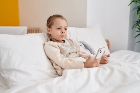 Foto de Adorable chica rubia usando teléfono inteligente sentado en la cama en el dormitorio - Imagen libre de derechos