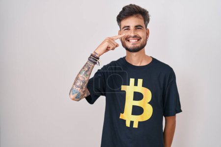Téléchargez les photos : Young hispanic man with tattoos wearing bitcoin t shirt pointing with hand finger to face and nose, smiling cheerful. beauty concept - en image libre de droit