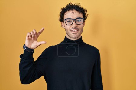 Téléchargez les photos : Hispanic man standing over yellow background smiling and confident gesturing with hand doing small size sign with fingers looking and the camera. measure concept. - en image libre de droit