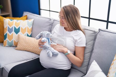 Photo for Young pregnant woman sitting on sofa holding rabbit doll at home - Royalty Free Image