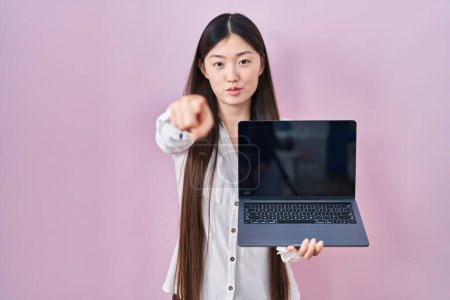 Photo for Chinese young woman holding laptop showing screen pointing with finger to the camera and to you, confident gesture looking serious - Royalty Free Image