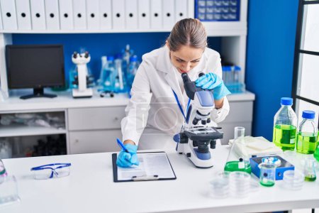 Photo for Young woman scientist using microscope writing on report at laboratory - Royalty Free Image