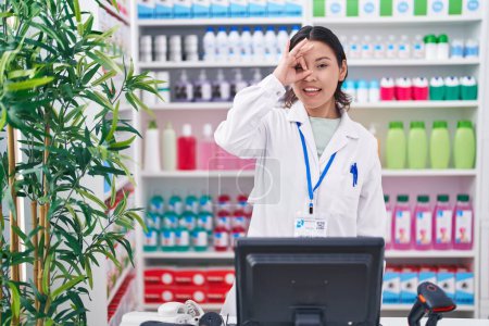 Téléchargez les photos : Jeune femme hispanique travaillant à la pharmacie pharmacie souriant heureux faire ok signe avec la main sur les yeux en regardant à travers les doigts - en image libre de droit