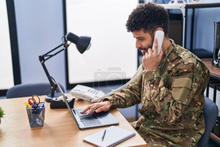 Téléchargez les photos : Jeune homme arabe soldat de l'armée utilisant un ordinateur portable parlant au téléphone au bureau - en image libre de droit