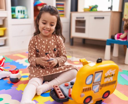 Foto de Adorable chica hispana sentada en el suelo jugando con un juguete mecánico en el jardín de infantes - Imagen libre de derechos