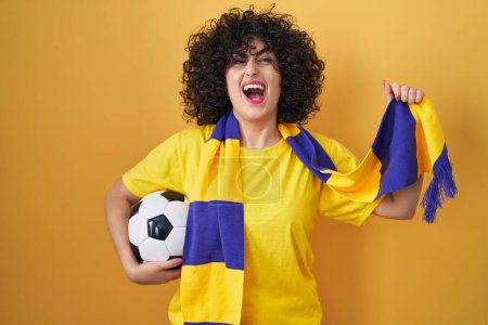 Photo for Young brunette woman with curly hair football hooligan holding ball angry and mad screaming frustrated and furious, shouting with anger looking up. - Royalty Free Image
