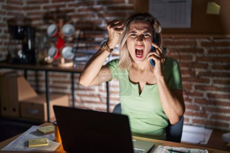 Foto de Young beautiful woman working at the office at night speaking on the phone angry and mad raising fist frustrated and furious while shouting with anger. rage and aggressive concept. - Imagen libre de derechos