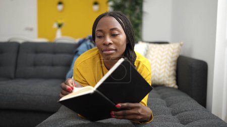 Téléchargez les photos : African woman reading book lying on sofa at home - en image libre de droit