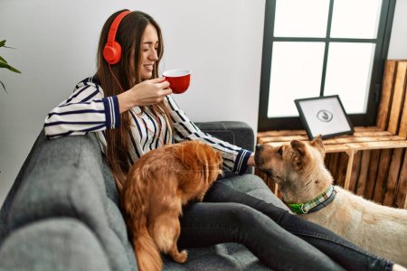 Téléchargez les photos : Young hispanic woman listening to music and drinking coffee sitting on sofa with dogs at home - en image libre de droit
