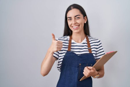 Photo for Young brunette woman wearing professional waitress apron and clipboard smiling happy and positive, thumb up doing excellent and approval sign - Royalty Free Image