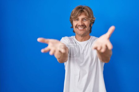 Photo for Middle age man standing over blue background smiling cheerful offering hands giving assistance and acceptance. - Royalty Free Image