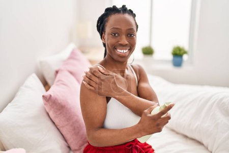 Téléchargez les photos : Femme afro-américaine appliquant un traitement de la peau assis sur le lit dans la chambre - en image libre de droit