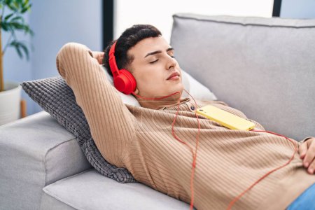 Téléchargez les photos : Jeune homme non binaire écoutant de la musique détendue sur le canapé à la maison - en image libre de droit