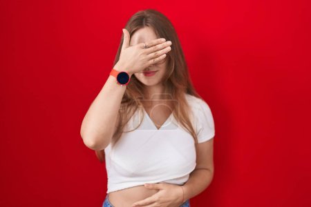 Photo for Young caucasian woman standing over red background covering eyes with hand, looking serious and sad. sightless, hiding and rejection concept - Royalty Free Image