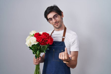 Foto de Young hispanic man holding bouquet of white and red roses beckoning come here gesture with hand inviting welcoming happy and smiling - Imagen libre de derechos