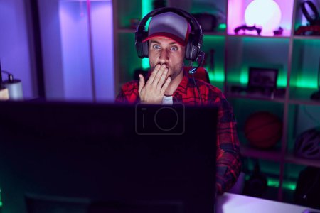 Téléchargez les photos : Jeune homme caucasien jouant à des jeux vidéo couvrant la bouche avec la main, choqué et effrayé par l'erreur. expression surprise - en image libre de droit