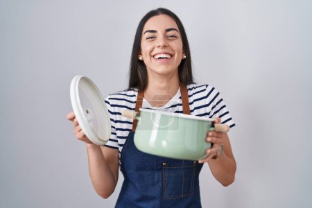 Photo for Young brunette woman wearing apron holding cooking pot smiling and laughing hard out loud because funny crazy joke. - Royalty Free Image