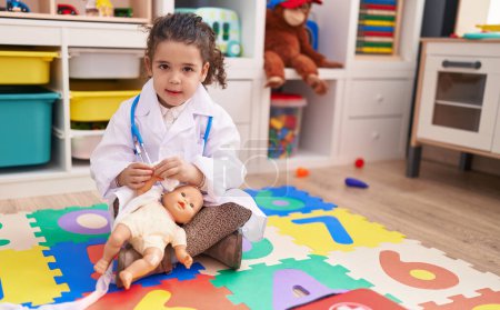 Photo for Adorable hispanic girl wearing doctor uniform bandaging arm baby doll at kindergarten - Royalty Free Image