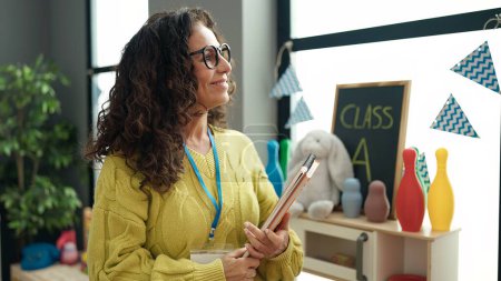 Téléchargez les photos : Moyen Age hispanique femme maternelle enseignant souriant confiant tenant des livres à la maternelle - en image libre de droit