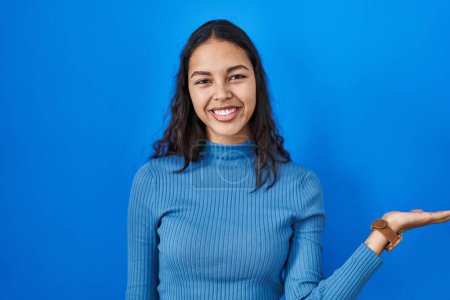 Foto de Young brazilian woman standing over blue isolated background smiling cheerful presenting and pointing with palm of hand looking at the camera. - Imagen libre de derechos
