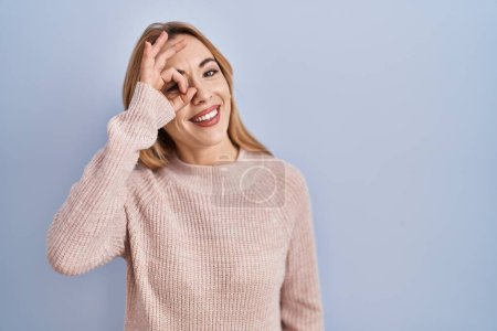 Photo for Hispanic woman standing over blue background doing ok gesture with hand smiling, eye looking through fingers with happy face. - Royalty Free Image