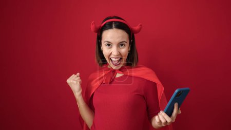 Téléchargez les photos : Young beautiful hispanic woman wearing devil costume using smartphone over isolated red background - en image libre de droit