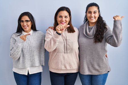 Photo for Mother and two daughters standing over blue background pointing to the back behind with hand and thumbs up, smiling confident - Royalty Free Image