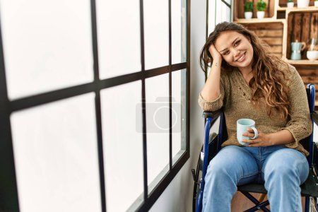 Téléchargez les photos : Jeune belle femme hispanique boire du café assis sur fauteuil roulant à la maison - en image libre de droit