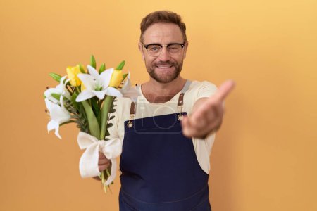 Foto de Middle age man with beard florist shop holding flowers smiling cheerful offering palm hand giving assistance and acceptance. - Imagen libre de derechos