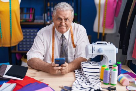 Téléchargez les photos : Homme moyen âge cheveux gris tailleur souriant confiant en utilisant smartphone au magasin de tailleur - en image libre de droit