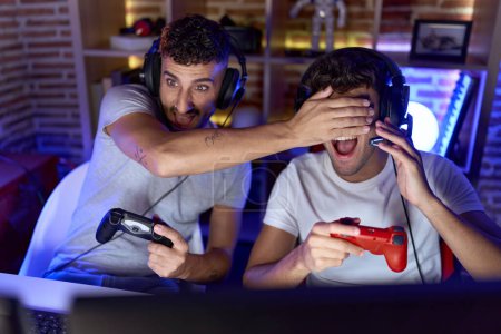 Foto de Two hispanic men streamers playing video game covering eyes cheating at gaming room - Imagen libre de derechos