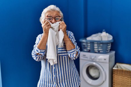 Foto de Senior grey-haired woman smiling confident smelling cleaning t shirt at laundry room - Imagen libre de derechos